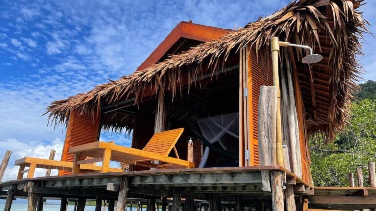 Un bungalow sobre pilotes con techo de palma, dos tumbonas de madera y una ducha exterior