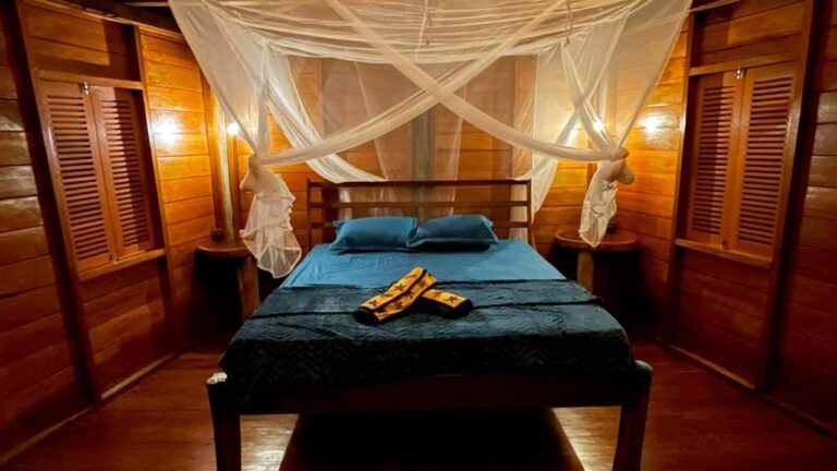 Una cama con sábanas azules y mosquitero, entre dos ventanas con contraventanas de madera