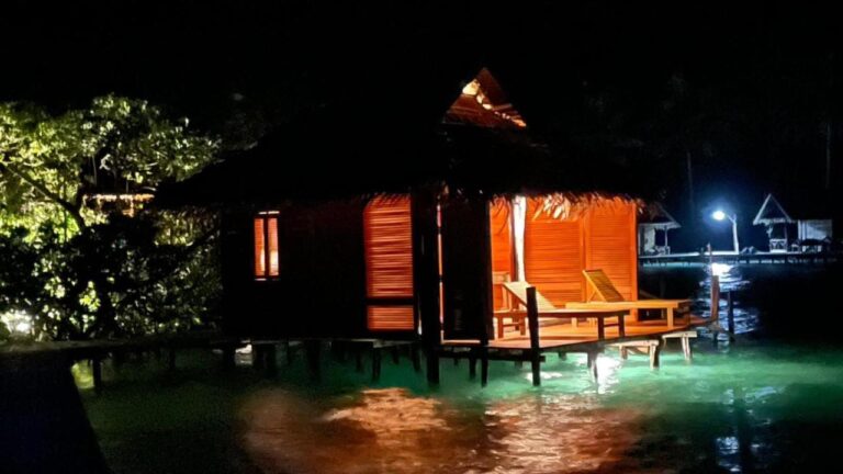 Un bungalow sobre pilotes iluminado de noche, con tumbonas en su terraza
