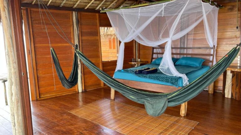 Una habitación de madera abierta al exterior, con una cama con sábanas azules y mosquitero y una hamaca verde