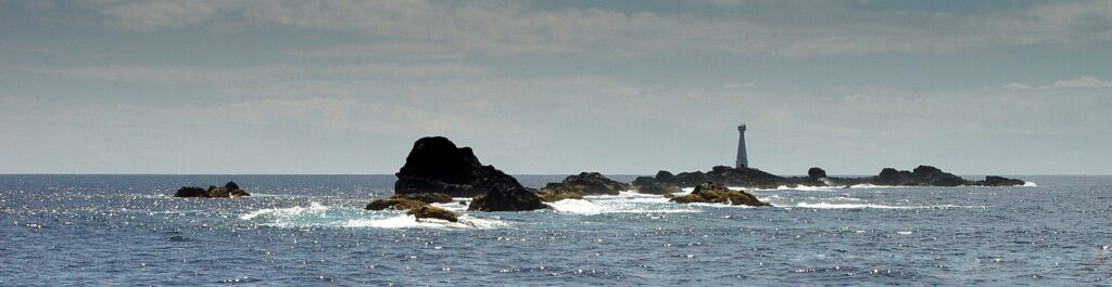 Panorámica de la cadena de escollos negros de las Formigas a ras del agua, ribeteados de espuma blanca sobre un mar azul brillante, con el faro blanco de Formigão a la derecha bajo un cielo velado