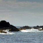 Panorámica de la cadena de escollos negros de las Formigas a ras del agua, ribeteados de espuma blanca sobre un mar azul brillante, con el faro blanco de Formigão a la derecha bajo un cielo velado