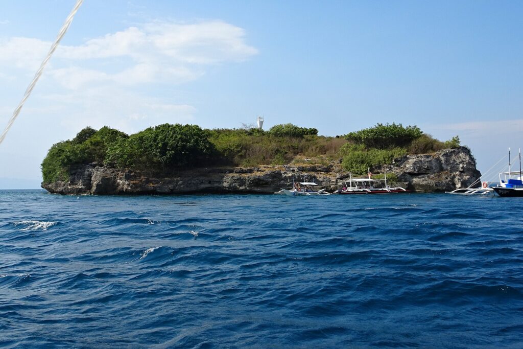 La isla Pescador vista desde el mar, cubierta de arbustos verdes, con su faro blanco en la cima y tres bancas de buceo amarradas al pie del acantilado