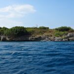 La isla Pescador vista desde el mar, cubierta de arbustos verdes, con su faro blanco en la cima y tres bancas de buceo amarradas al pie del acantilado