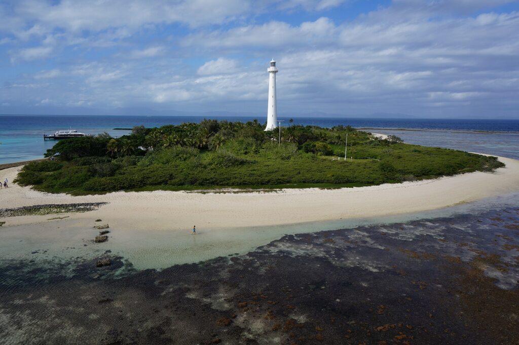 El gran faro blanco del islote Amédée se alza sobre la vegetación, con el islote rodeado de arena blanca, la plataforma de coral al descubierto en primer plano y un barco amarrado a la izquierda