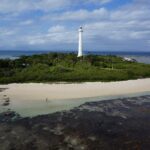 El gran faro blanco del islote Amédée se alza sobre la vegetación, con el islote rodeado de arena blanca, la plataforma de coral al descubierto en primer plano y un barco amarrado a la izquierda
