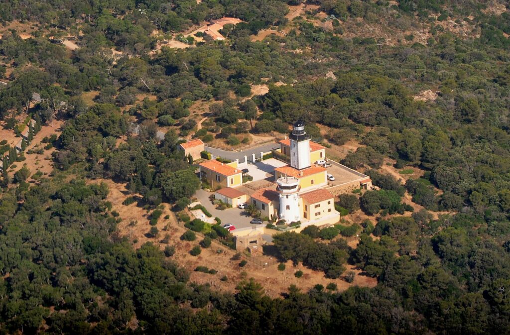 Vista aérea del faro del cabo Camarat, una torre blanca con linterna negra rodeada de edificios amarillos con tejados de teja naranja, en medio de un denso bosque mediterráneo.