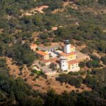 Vista aérea del faro del cabo Camarat, una torre blanca con linterna negra rodeada de edificios amarillos con tejados de teja naranja, en medio de un denso bosque mediterráneo.