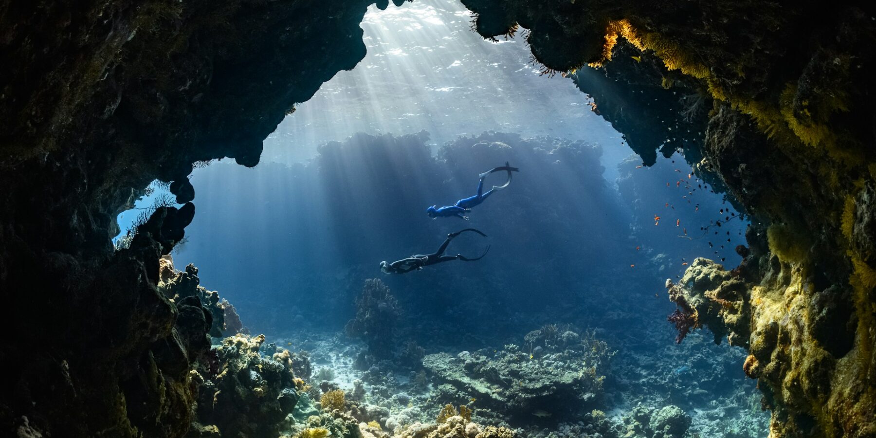 Dos buceadores atraviesan una cueva submarina iluminada por los rayos del sol, sobre un arrecife