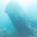 La popa rota del Thistlegorm, levantada e inclinada en el azul, con un buceador a la derecha