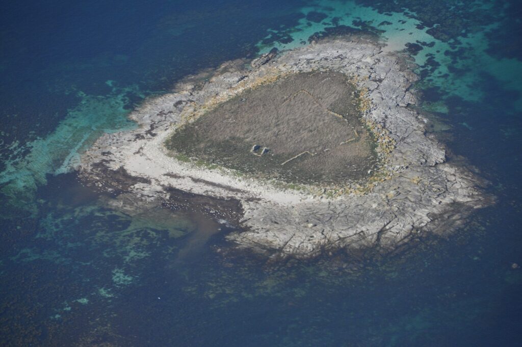 Vista aérea de un pequeño islote de losas de granito gris, cubierto en el centro de hierba seca donde se adivinan muretes en ruinas, bordeado de una playa clara y rodeado de bajos turquesa y de agua azul profundo.