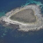 Vista aérea de un pequeño islote de losas de granito gris, cubierto en el centro de hierba seca donde se adivinan muretes en ruinas, bordeado de una playa clara y rodeado de bajos turquesa y de agua azul profundo.