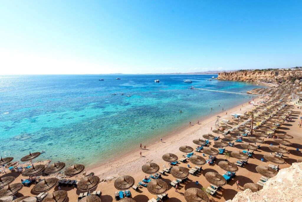 Una larga playa de arena al pie de un acantilado, con sombrillas de paja y un pontón sobre el mar turquesa