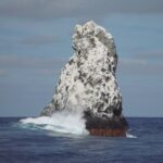 El pico noroeste de Roca Partida, blanco de guano, visto desde el barco, con una ola que rompe en su base y aves en vuelo encima
