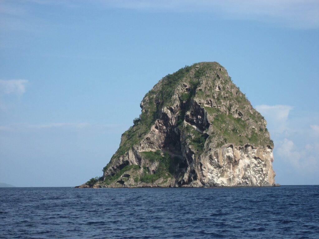 La Roca del Diamante vista de cerca desde el mar, una masa de roca gris horadada de cuevas, con vetas blancas y coronada de vegetación, sobre un mar azul oscuro