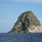 La Roca del Diamante vista de cerca desde el mar, una masa de roca gris horadada de cuevas, con vetas blancas y coronada de vegetación, sobre un mar azul oscuro