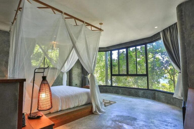 Una habitación con paredes de hormigón, una cama con mosquitero y un ventanal que da a los árboles
