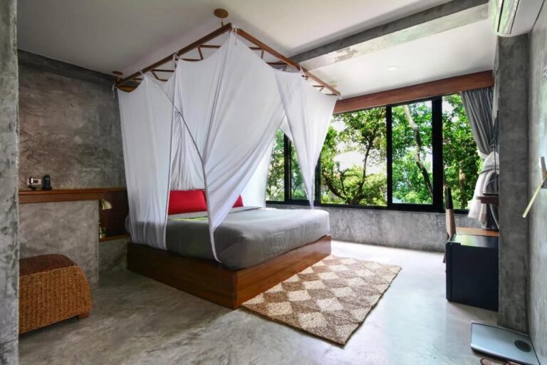 Una cama con mosquitero y cojines rojos, frente a una gran ventana que da al bosque