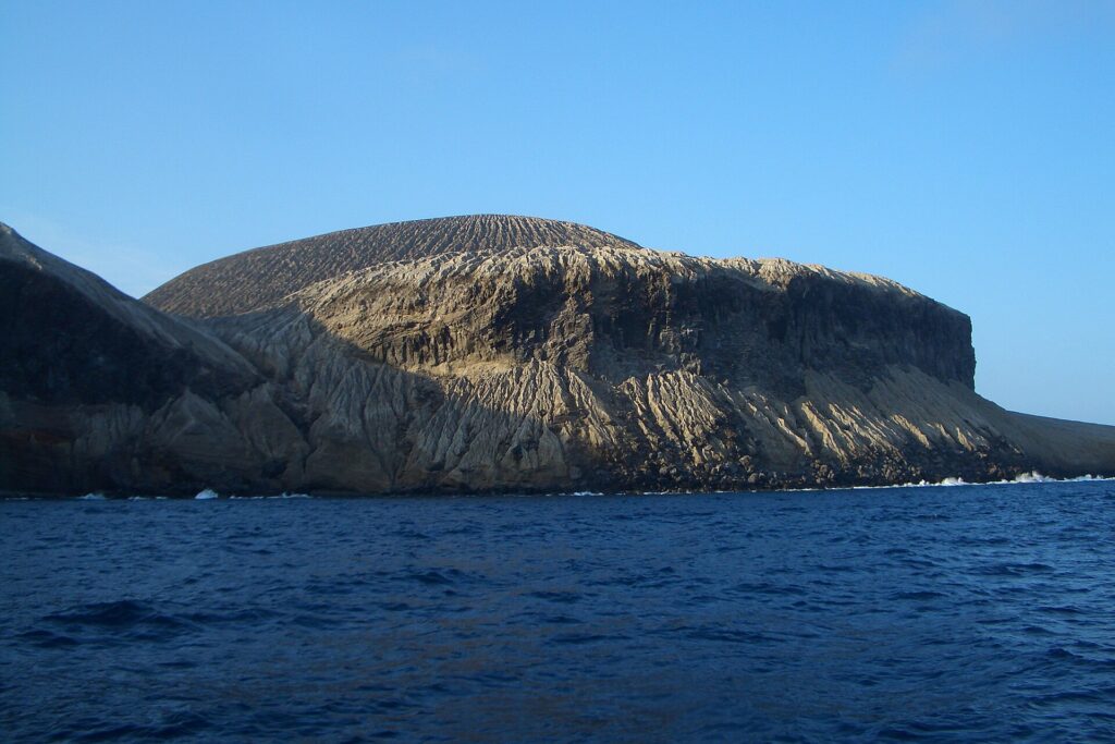 La costa de San Benedicto vista desde el mar: el cono de ceniza del volcán Bárcena, surcado de barrancos, sobre acantilados oscuros y un mar azul oscuro