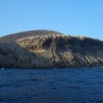 La costa de San Benedicto vista desde el mar: el cono de ceniza del volcán Bárcena, surcado de barrancos, sobre acantilados oscuros y un mar azul oscuro