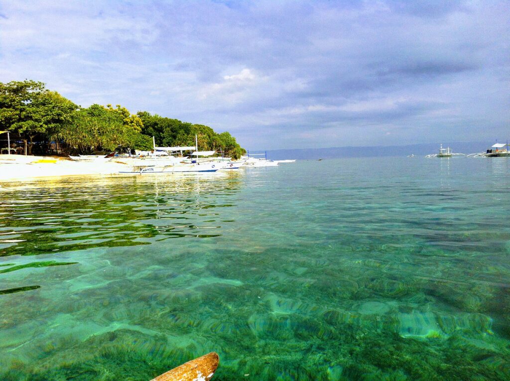Agua turquesa transparente sobre un fondo poco profundo, frente a una playa de arena blanca bordeada de árboles donde se alinean bangkas blancas, con otros barcos mar adentro bajo un cielo nublado.