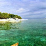 Agua turquesa transparente sobre un fondo poco profundo, frente a una playa de arena blanca bordeada de árboles donde se alinean bangkas blancas, con otros barcos mar adentro bajo un cielo nublado.