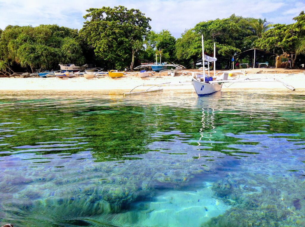 A ras del agua, cabezas de coral bajo un agua clara y turquesa, frente a una playa de arena blanca donde fondea una bangka blanca, con otras barcas de colores varadas bajo los árboles.
