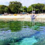 A ras del agua, cabezas de coral bajo un agua clara y turquesa, frente a una playa de arena blanca donde fondea una bangka blanca, con otras barcas de colores varadas bajo los árboles.