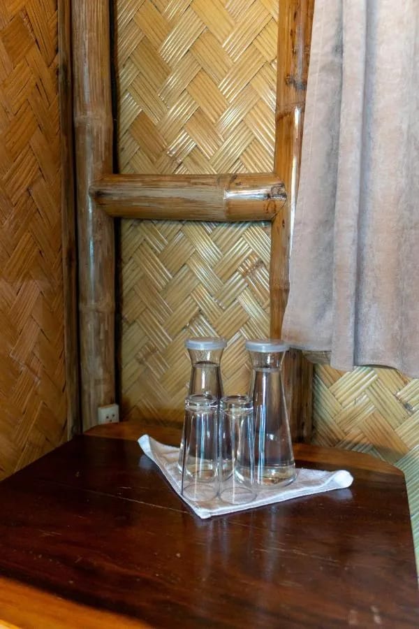 Dos jarras de agua y vasos sobre una mesa de madera, junto a una pared de bambú trenzado