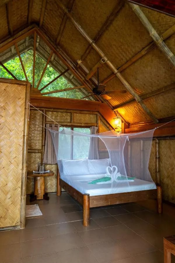 Una habitación bajo un alto techo de bambú trenzado, con una cama con mosquitero y un ventilador de techo