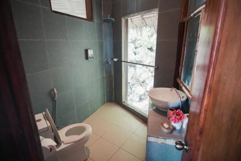 Un baño con azulejos verde oscuro, con lavabo, ducha y una ventana que da a las rocas