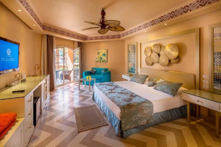 Una habitación con cama grande, ventilador de techo, un sillón azul y suelo en espiga