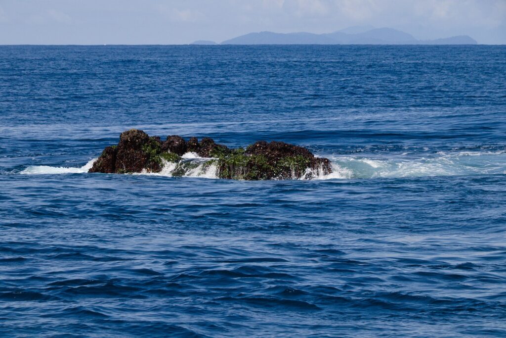 La cima de Richelieu Rock asoma con la marea baja, una roca oscura cubierta de algas verdes y batida por las olas, con las islas Surin en el horizonte
