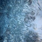 Un banco cerrado de sardinas plateadas roza un fondo de arena muy clara salpicado de rocas oscuras, con la masa de peces más densa hacia la izquierda, en el azul.