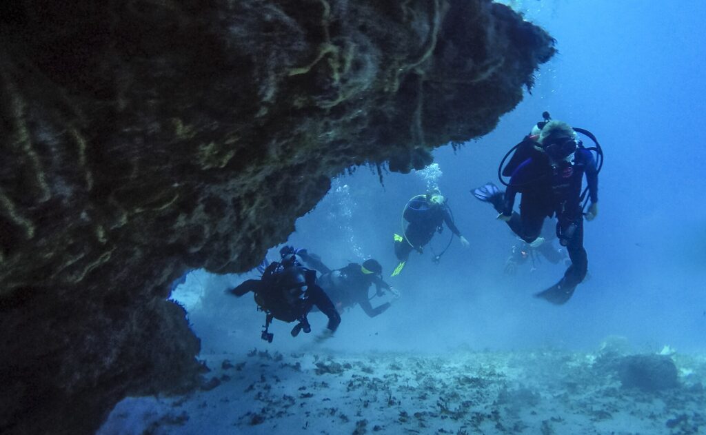 Cuatro buceadores salen de un túnel de coral sobre un fondo de arena, en Cozumel, con el techo rocoso sobre ellos