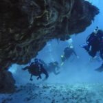 Cuatro buceadores salen de un túnel de coral sobre un fondo de arena, en Cozumel, con el techo rocoso sobre ellos