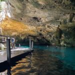 El ojo este del cenote Dos Ojos: una bóveda de caliza erizada de estalactitas sobre un agua turquesa, con un pontón de madera a la izquierda y bañistas al fondo
