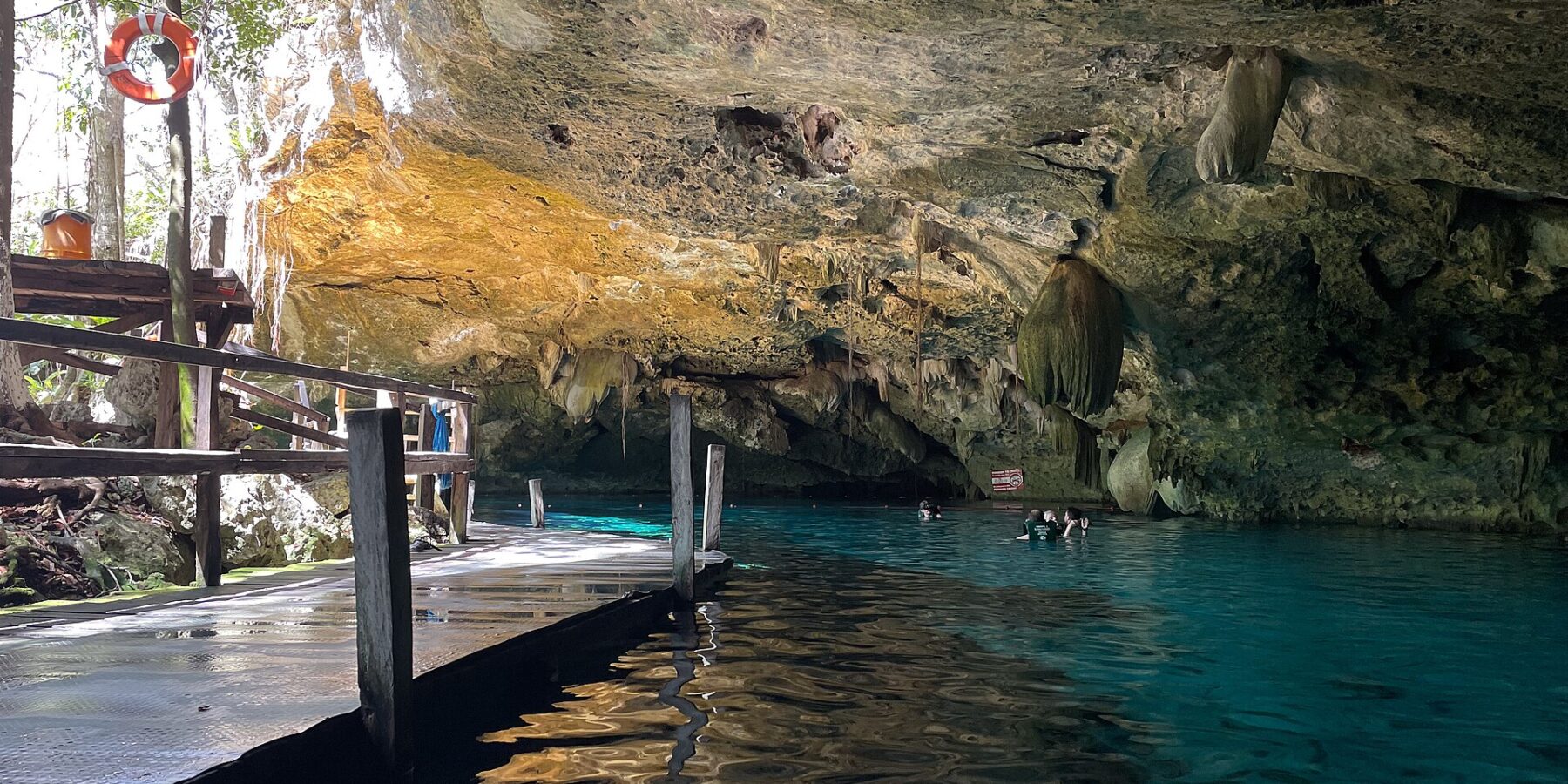 El ojo este del cenote Dos Ojos: una bóveda de caliza erizada de estalactitas sobre un agua turquesa, con un pontón de madera a la izquierda y bañistas al fondo