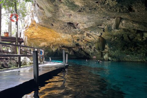 El ojo este del cenote Dos Ojos: una bóveda de caliza erizada de estalactitas sobre un agua turquesa, con un pontón de madera a la izquierda y bañistas al fondo
