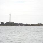 Un faro blanco y esbelto con la puerta roja se alza sobre el islote bajo y rocoso de Formigão, de rocas oscuras bordeadas de algas pardas, sobre un mar gris y agitado bajo un cielo cubierto