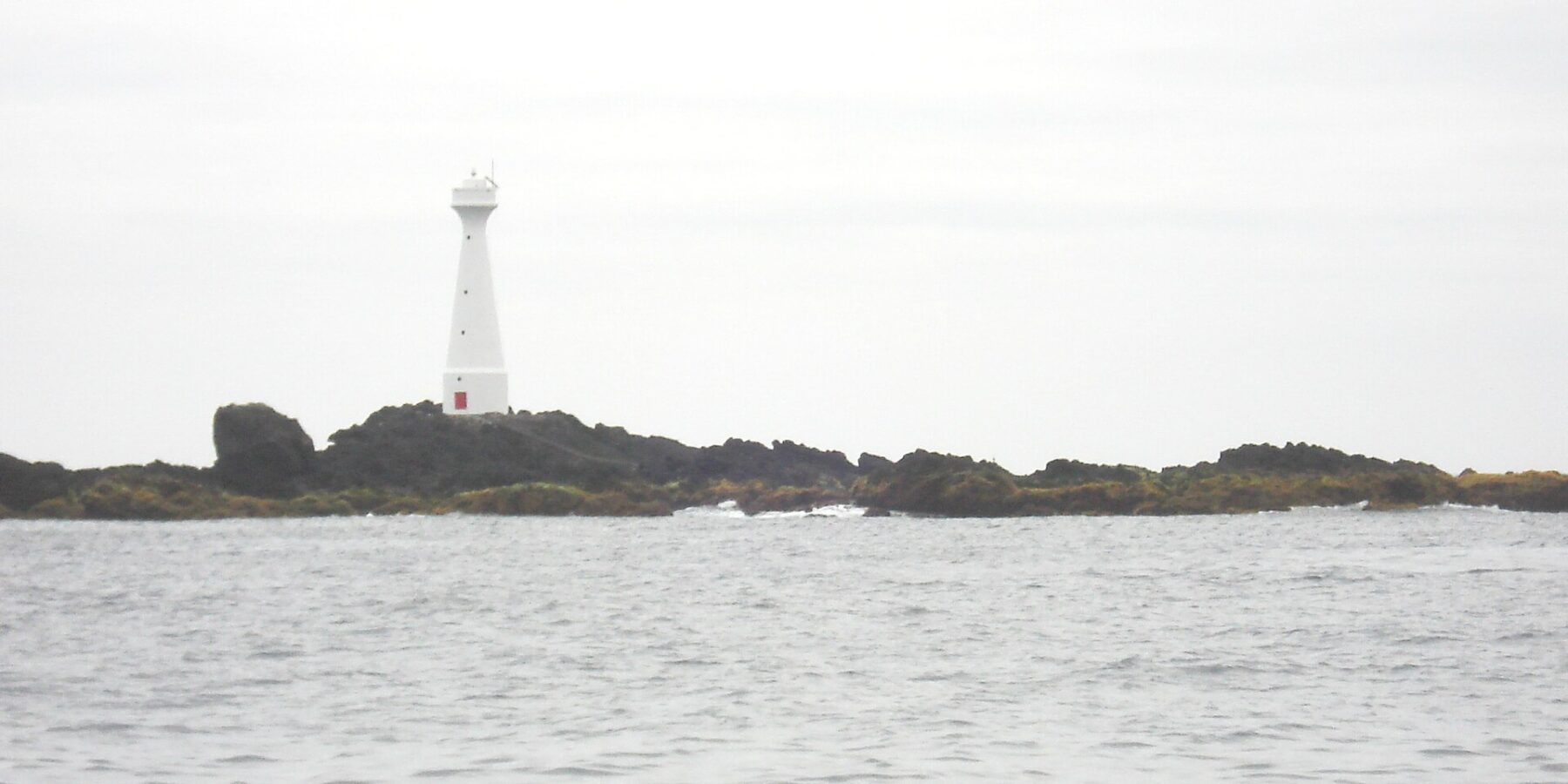 Un faro blanco y esbelto con la puerta roja se alza sobre el islote bajo y rocoso de Formigão, de rocas oscuras bordeadas de algas pardas, sobre un mar gris y agitado bajo un cielo cubierto
