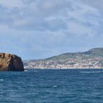 Panorámica de los dos islotes de toba ocre de Madalena sobre un mar azul oscuro, el Ilhéu Deitado largo y plano a la izquierda y el Ilhéu em Pé erguido a la derecha, con la ciudad blanca de Horta y las colinas verdes de Faial al fondo