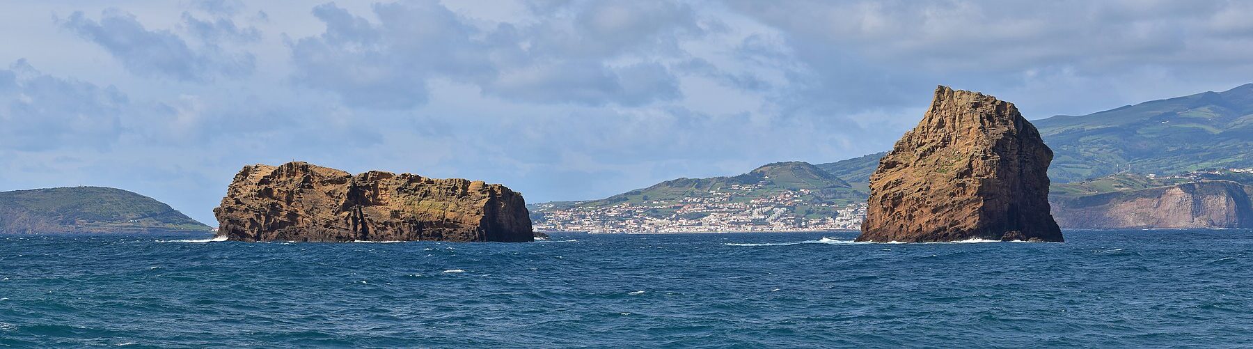 Panorámica de los dos islotes de toba ocre de Madalena sobre un mar azul oscuro, el Ilhéu Deitado largo y plano a la izquierda y el Ilhéu em Pé erguido a la derecha, con la ciudad blanca de Horta y las colinas verdes de Faial al fondo