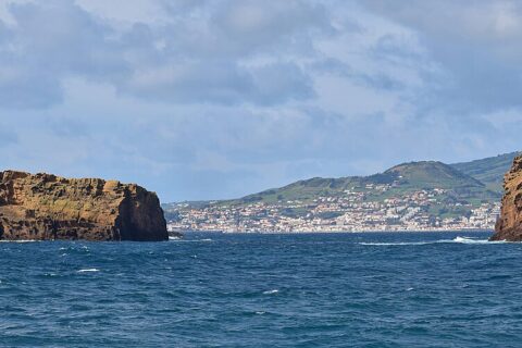 Panorámica de los dos islotes de toba ocre de Madalena sobre un mar azul oscuro, el Ilhéu Deitado largo y plano a la izquierda y el Ilhéu em Pé erguido a la derecha, con la ciudad blanca de Horta y las colinas verdes de Faial al fondo