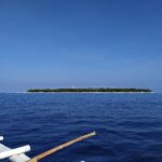 La isla llana y arbolada de Balicasag en el horizonte, con su faro blanco y su franja de arena clara, vista desde una bangka cuyos balancines blancos ocupan el primer plano, bajo un cielo azul sin nubes.
