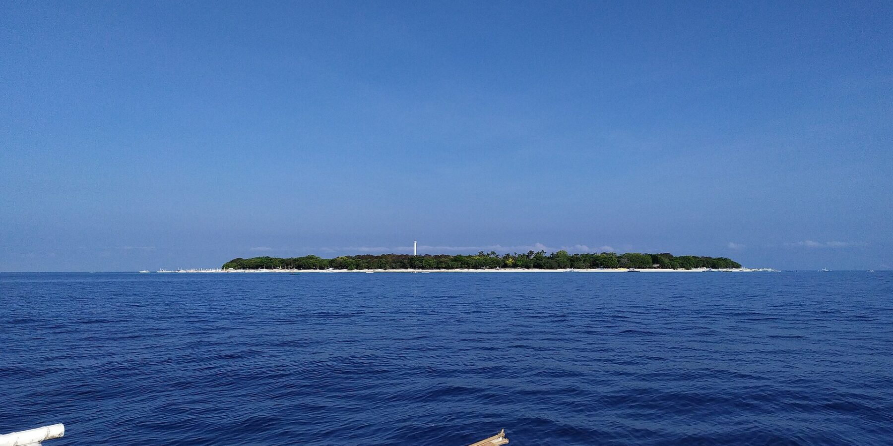 La isla llana y arbolada de Balicasag en el horizonte, con su faro blanco y su franja de arena clara, vista desde una bangka cuyos balancines blancos ocupan el primer plano, bajo un cielo azul sin nubes.