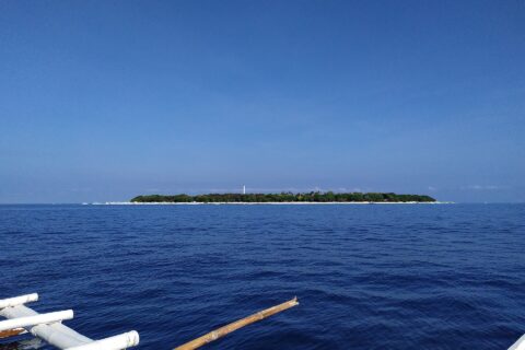 La isla llana y arbolada de Balicasag en el horizonte, con su faro blanco y su franja de arena clara, vista desde una bangka cuyos balancines blancos ocupan el primer plano, bajo un cielo azul sin nubes.