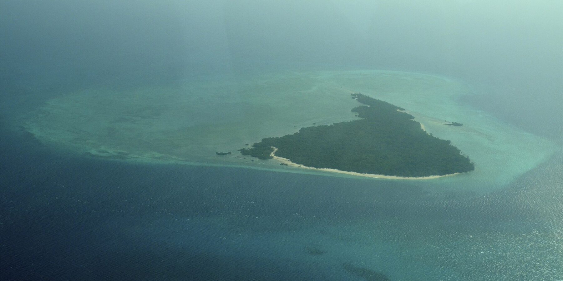 La isla Misali vista desde el avión, un triángulo de bosque bordeado por una playa blanca, sobre una plataforma arrecifal turquesa en medio de un mar azul oscuro, entre la bruma