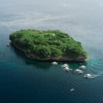 Vista aérea de la isla Pescador, una roca cubierta de vegetación verde con su faro blanco, rodeada de una decena de bancas de buceo sobre un mar azul oscuro