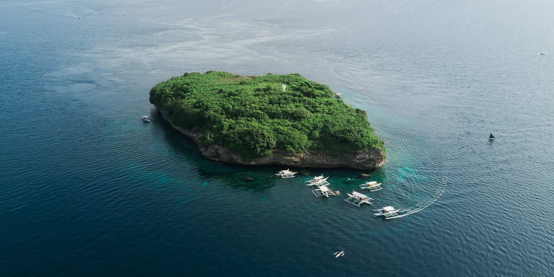 Vista aérea de la isla Pescador, una roca cubierta de vegetación verde con su faro blanco, rodeada de una decena de bancas de buceo sobre un mar azul oscuro